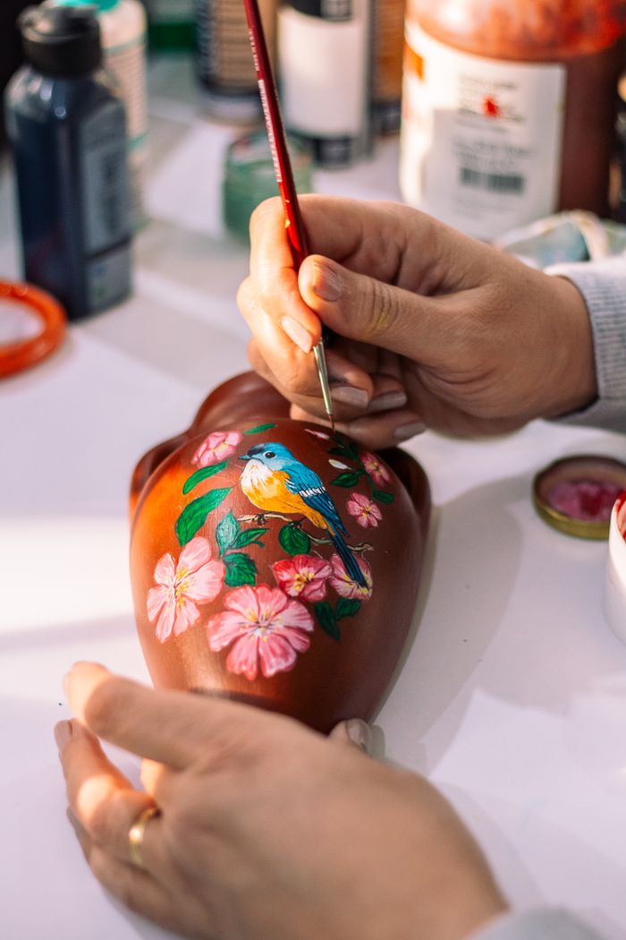 Close-up of hands painting a bird and flowers on a ceramic vase.