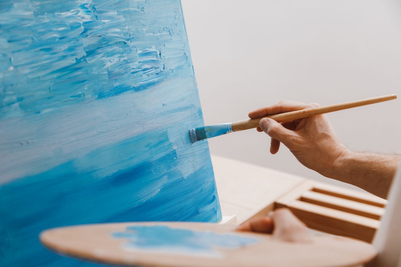 A close-up image of an artist using a brush to paint with shades of blue on a canvas indoors.